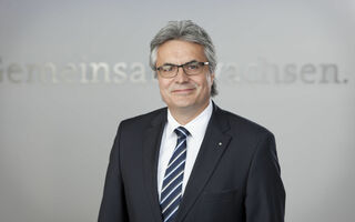 Portrait eines Mannes mittleren Alters mit grauen Haaren und heller Hautfarbe. Er trägt eine Brille und einen Anzug.