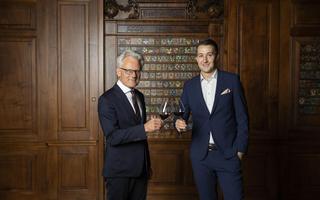 Zwei Männer in Anzügen stehen vor einer dunkelbraunen Holzwand und stossen mit einem Glas Rotwein an.