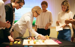 Mit Hilfe eines physischen Holzspielbretts mit einer eingravierten Matrix und dem Einsatz von verschiedenen Bausteinen wird die Strategie greifbar.