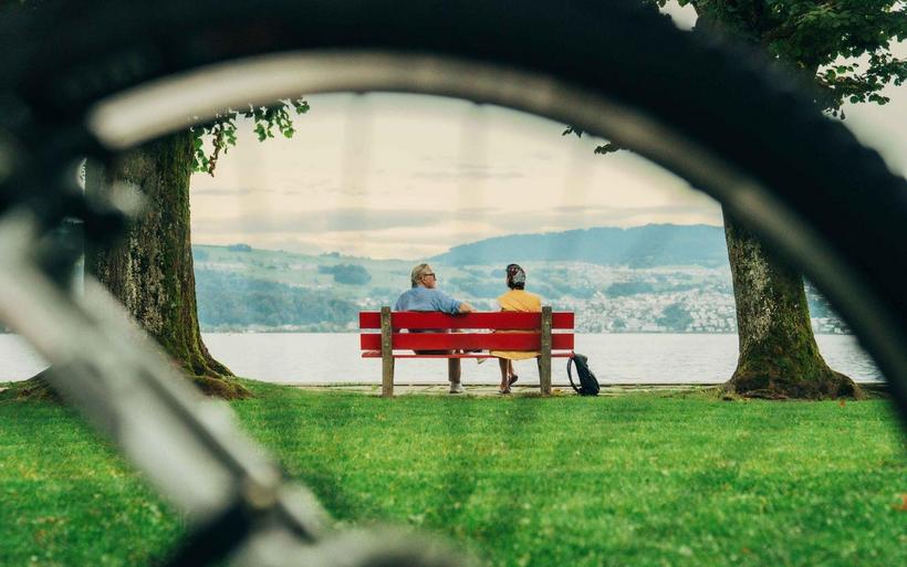 Zwei Personen sitzen auf einer roten Bank am See hinter grüner Wiese